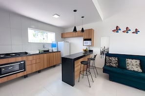 A modern kitchen with a central island, wooden cabinets, and a dining area with a teal sofa and bar stools. - Apartamento 3 com 2 suítes 450m da praia (Santa Cruz Cabrália)