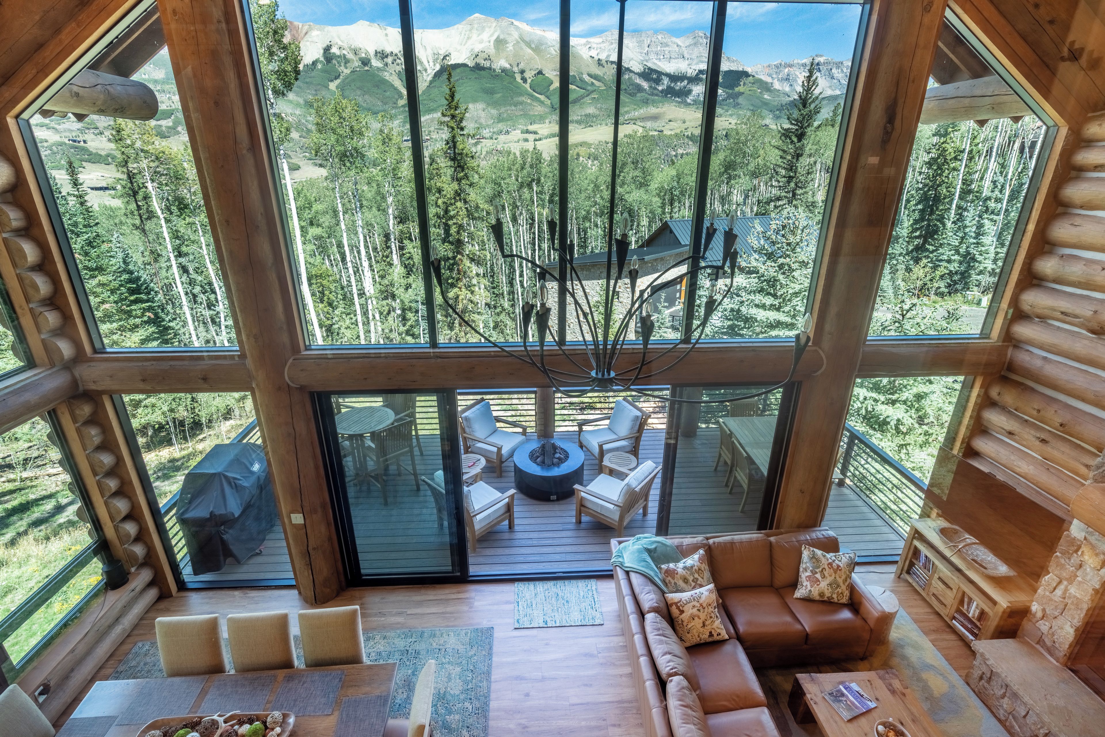 A log cabin with a large glass window offering a mountain view. Inside, there is a wooden deck with outdoor furniture and a fireplace.