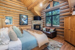 Interior de una cabaña de madera con una cama, chimenea, sofá y vista al bosque a través de amplias ventanas.