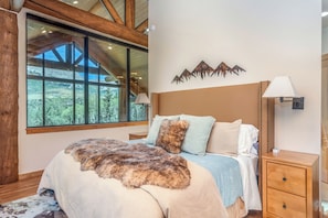 A bedroom with a large window, wooden beams, a bed with a fur throw, and a mountain art piece on the wall.