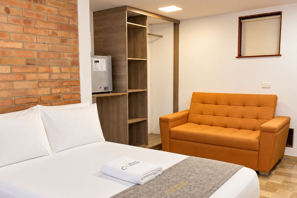 A hotel room with a bed, a grey textured throw, a white towel, an orange armchair, and a wooden wardrobe.