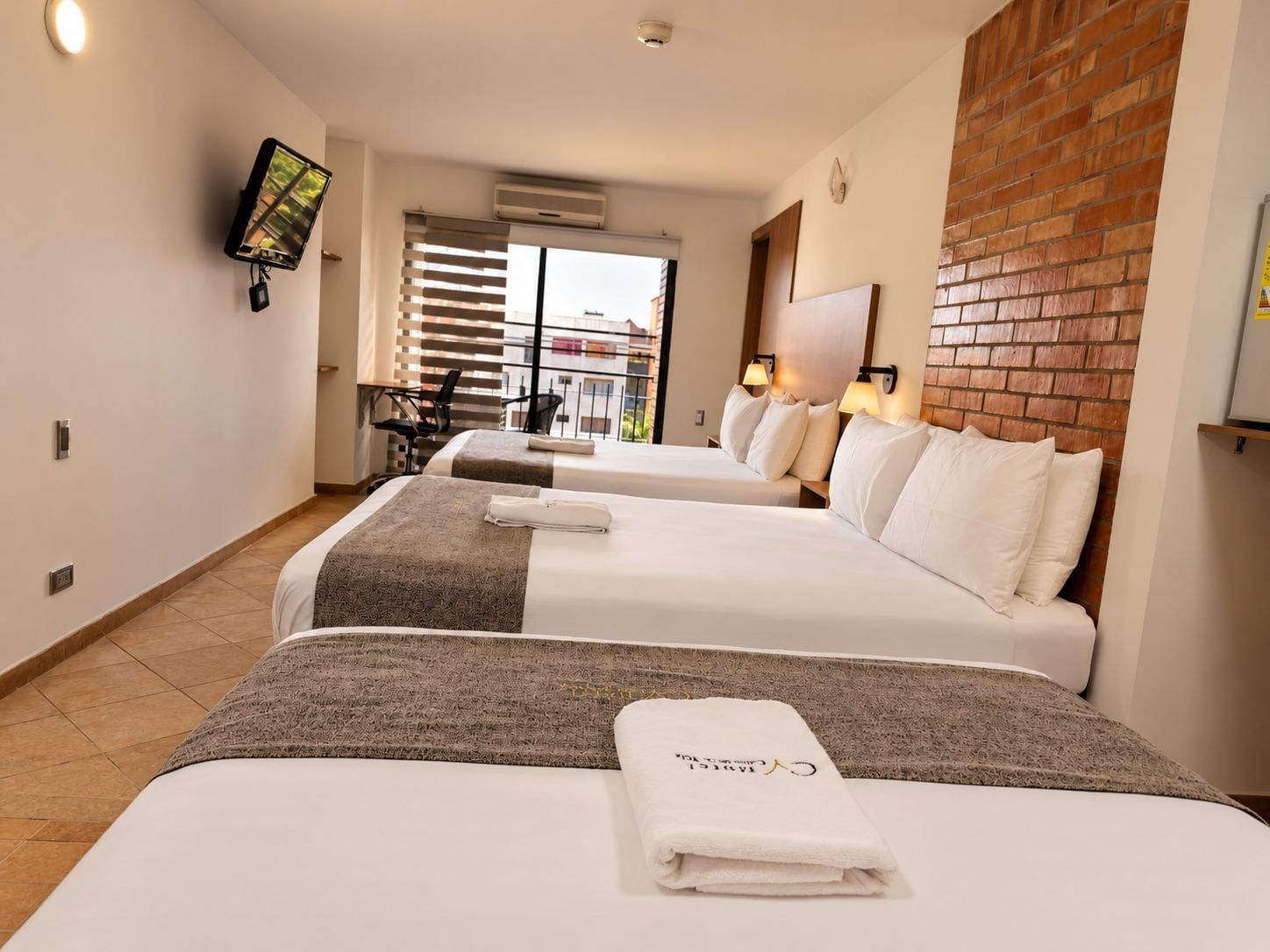 Habitación de hotel con dos camas, un televisor montado en la pared, un muro decorativo de ladrillo y vista al exterior.