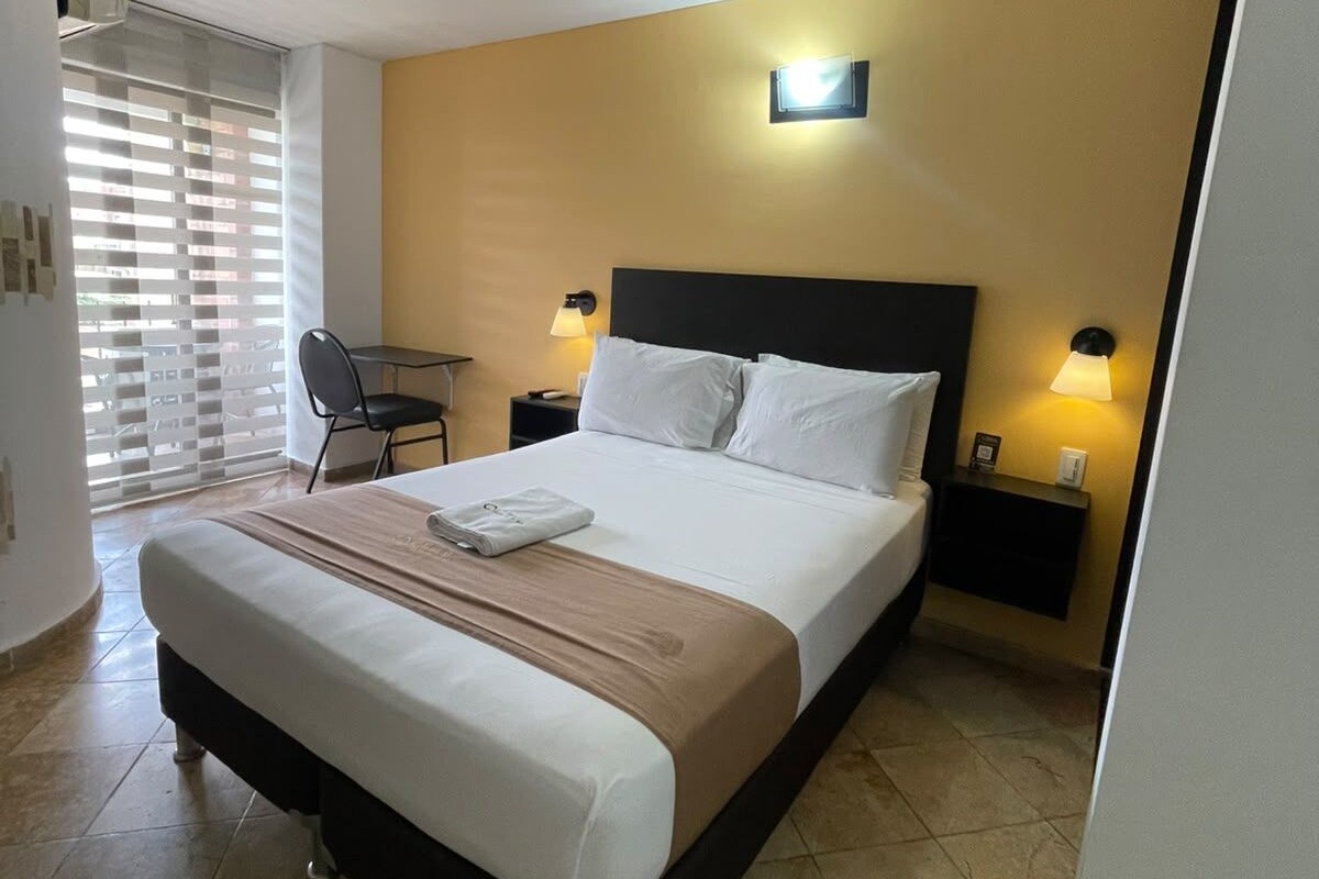 A hotel room with a bed, bedside tables, a chair, and a window with blinds.