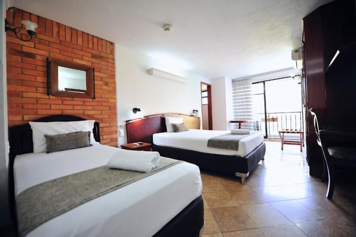Habitació d'hotel amb dos llits, una paret d'accent de maó, una finestra petita i un balcó.
