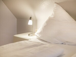 Un lit bien fait, avec une literie blanche, dans une pièce dont le plafond est en pente et qui est équipée d’une table de chevet avec une lampe.