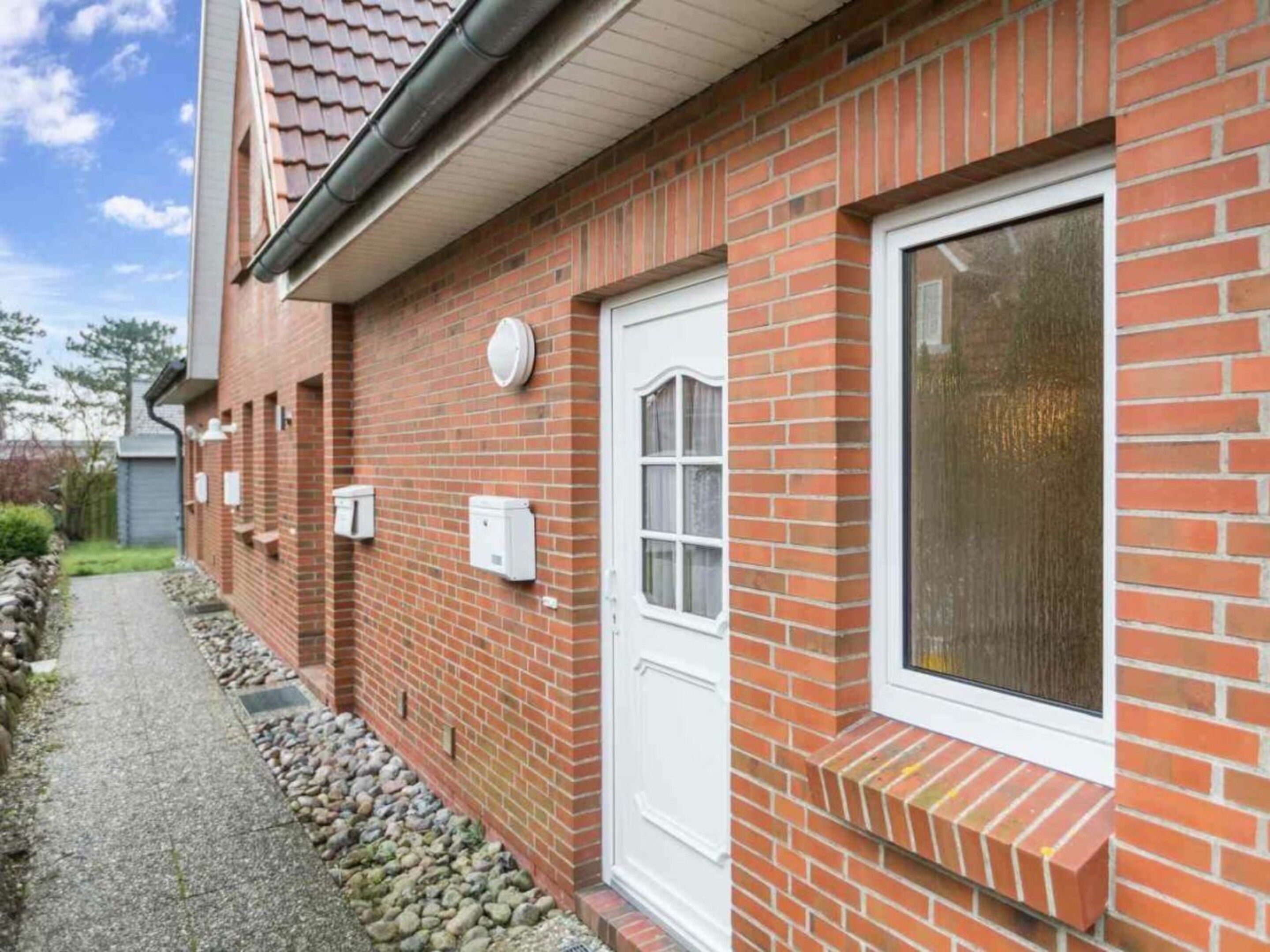 A brick building with a white door and window, a gravel path, and a small garden area.