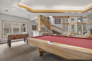Ein Billardtisch in einem Raum mit einer Holztreppe, einem Kicker und Blick durch große Fenster.