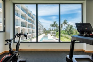 A room with a large window overlooking a pool and a building with balconies. There is a treadmill and an exercise bike inside. - Born | 2-bedroom modern apartment with pools (Bávaro)