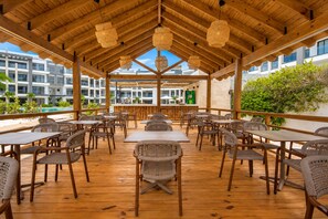 An outdoor dining area with wooden tables and chairs, a bar, and a view of buildings and greenery. - Born | 2-bedroom modern apartment with pools (Bávaro)