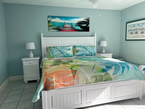 A bedroom with a white bed, two bedside tables, a nightstand, a painting of a beach scene, and a framed picture on the wall. - Runaway Bay 103 - Beach, Pool and activities galore (Bradenton Beach)