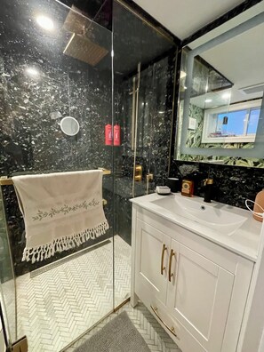 Ein modernes Badezimmer mit einer gläsernen Duschkabine, einem weißen Waschtisch mit goldenen Griffen und einem großen Spiegel, der den Raum reflektiert.