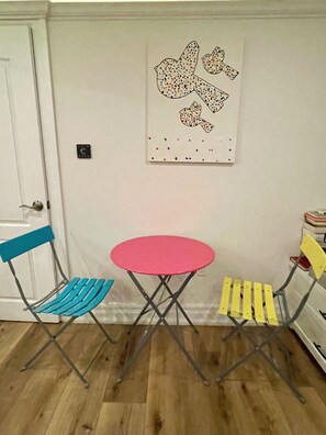 Ein Zimmer mit Holzboden, einer weißen Tür, einem rosafarbenen Tisch und zwei Stühlen mit blau-gelben Rückenlehnen.