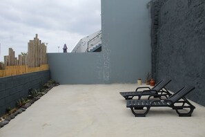 Un patio de concreto con dos sillones reclinables negros, cerca de madera y una pared gris con una escalera metálica.