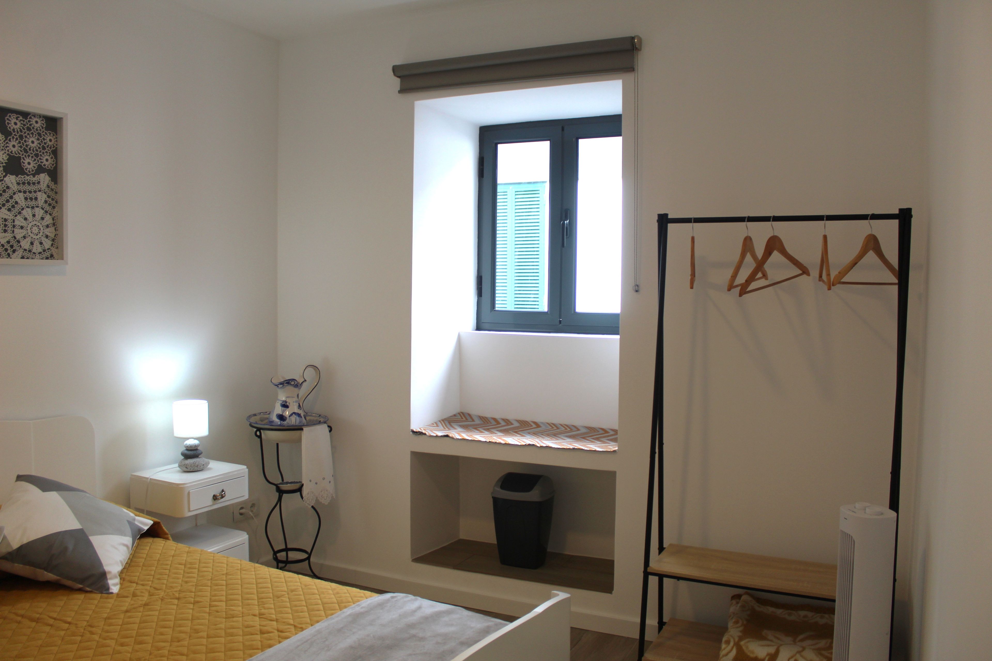 Un dormitorio con cama, mesita de noche, lámpara, ventana y perchero.
