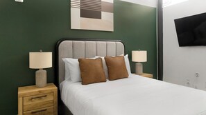 Habitación de hotel con una cama, dos mesitas de noche, un televisor montado en la pared y un cuadro sobre la cama.