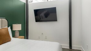 Habitación de hotel con cama, mesita de noche con lámpara, televisor montado en la pared y una pared de acento verde.