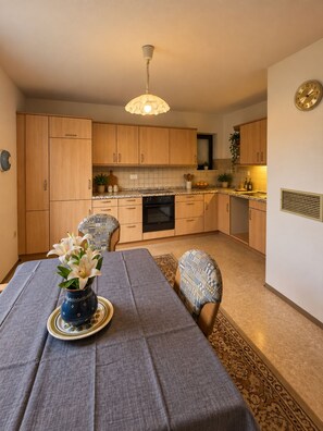 A dining area with a table set for two, a kitchen in the background, and a ceiling light fixture. - Romantic, Renovated Baierweiher Hunting Lodge in Neubäu am See (Roding)