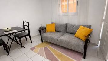 Una sala de estar con un sofá gris, dos cojines amarillos, una mesa de comedor con sillas y un ventanal con cortinas.
