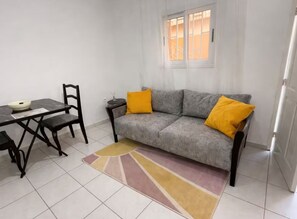 A living room with a grey sofa, two yellow cushions, a dining table with chairs, and a window with curtains. - Ideal apartment with bedroom, living room, kitchen, and shower (louis)