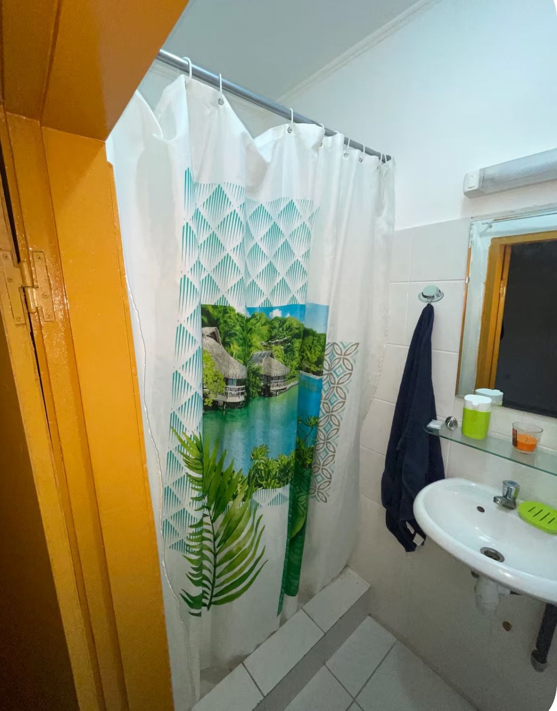 A bathroom with a shower curtain featuring a tropical landscape design, a white sink with a green soap dish, and a mirror above the sink.