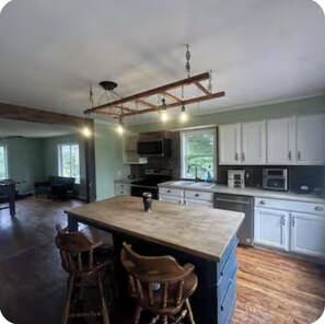 A spacious kitchen with a large island, wooden chairs, and a hanging light fixture. - Rustic 1904 Estate on a Working WI Wedding Venue - Sleeps 16 (Sparta)