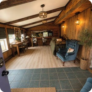 A rustic interior with a wooden ceiling, a blue armchair, a dining area, and a bar. - Rustic 1904 Estate on a Working WI Wedding Venue - Sleeps 16 (Sparta)