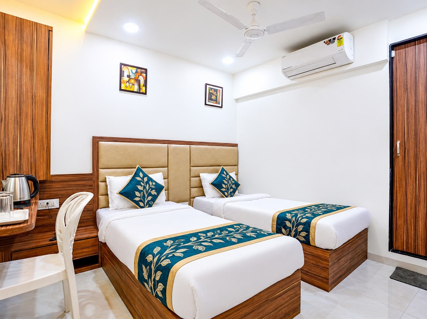 A hotel room with two beds, a desk with a kettle, a chair, and an air conditioning unit.