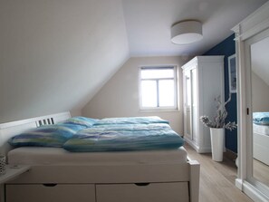 Ein Schlafzimmer mit schrägverlaufender Decke, einem Bett mit blau-weiß gestreifter Bettdecke, einem weißen Nachttisch, einer Zimmerpflanze und einem Kleiderschrank.