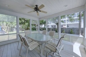 A covered outdoor dining area with a round table and six chairs, a ceiling fan, and large windows offering views of a residential area. - Coastal Charm Near the Beach in Rehoboth (Rehoboth Beach)