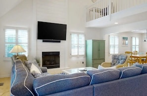 A spacious living room with a large sofa, a flat-screen TV mounted on the wall, a fireplace, and a dining area with a table and chairs. - Coastal Charm Near the Beach in Rehoboth (Rehoboth Beach)