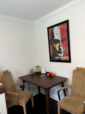 Een eetgedeelte met een donker houten tafel, twee stoelen, een ingelijst kunstwerk, een klein uiltje, een bord en een schaal met fruit.
