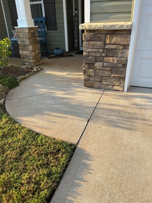 A concrete driveway leading to a stone pillar with a 'WELCOME' sign, a stone wall, and a door.