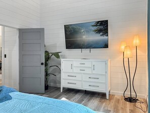 Quarto com cama, cômoda branca, televisão fixada na parede e abajur.
