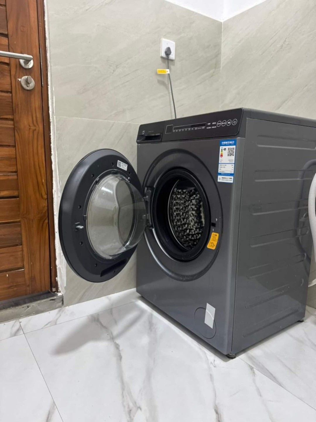 Eine Waschmaschine mit Frontöffnung in einem Badezimmer, mit Holztür und gefliesten Wänden.