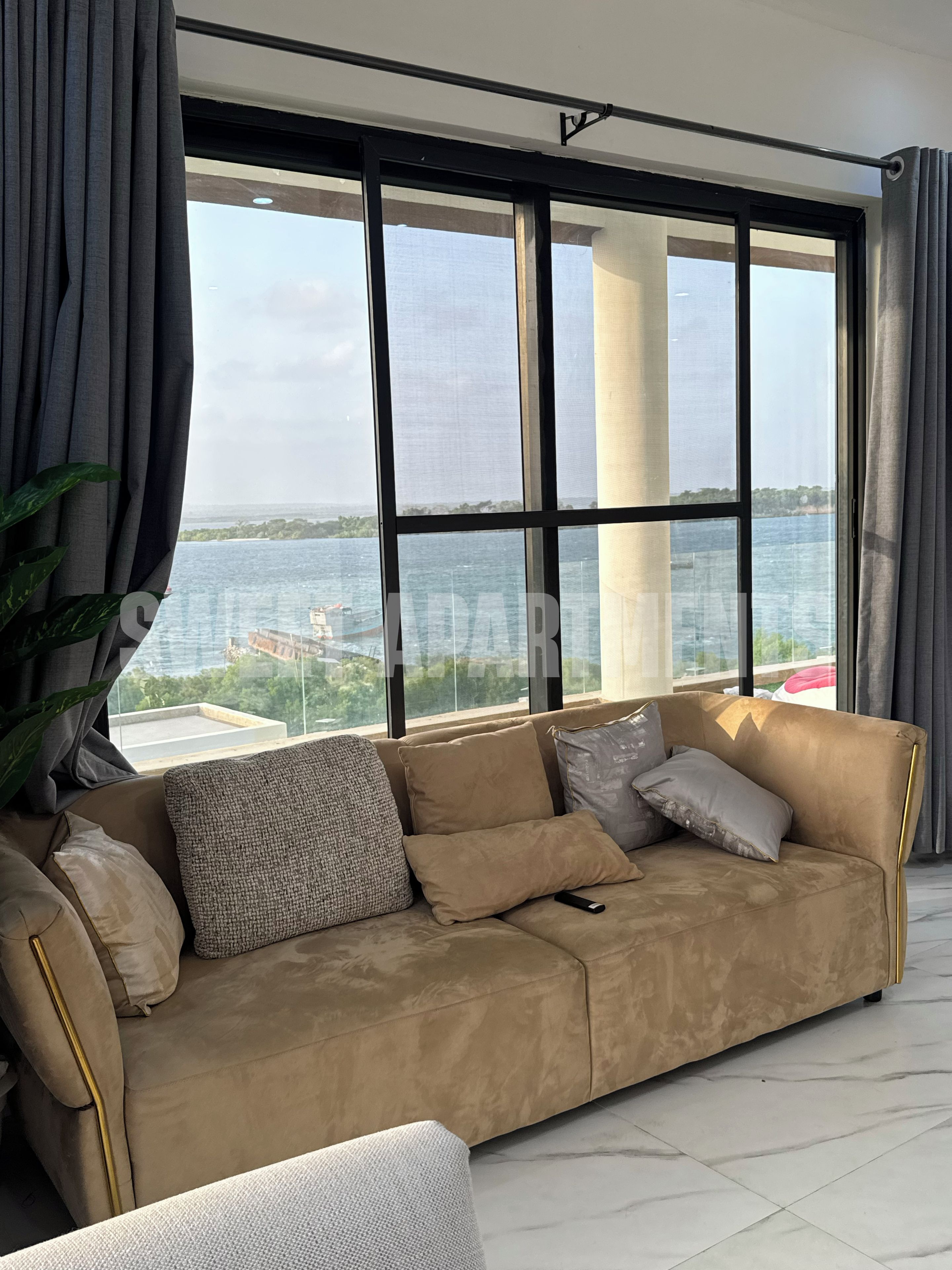 Un salón moderno con un sofá color beige, un ventanal con vistas al mar y una planta en maceta.
