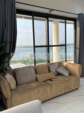 A modern living room with a beige sofa, a large window with a view of the sea, and a potted plant.
