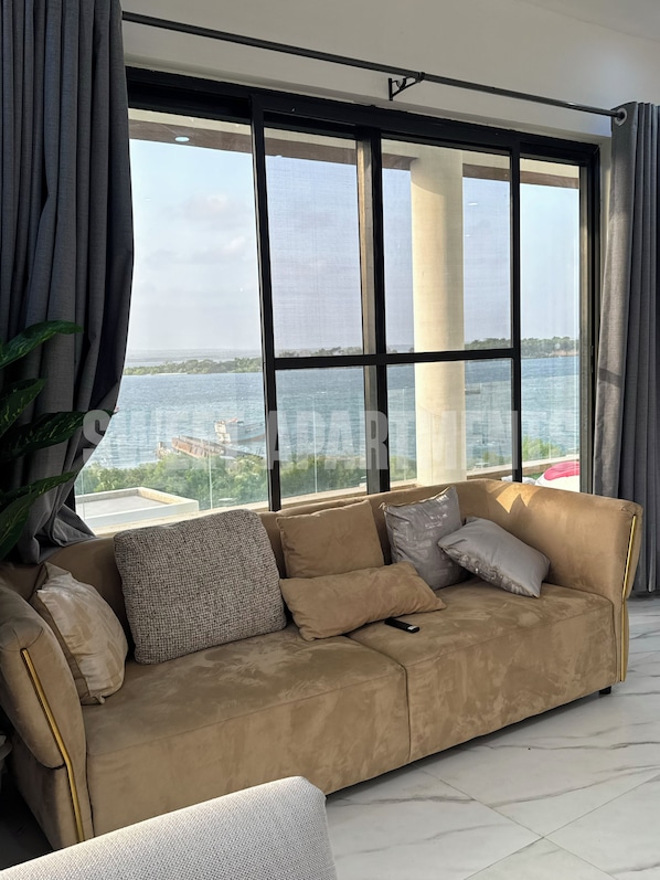 A modern living room with a beige sofa, a large window with a view of the sea, and a potted plant.