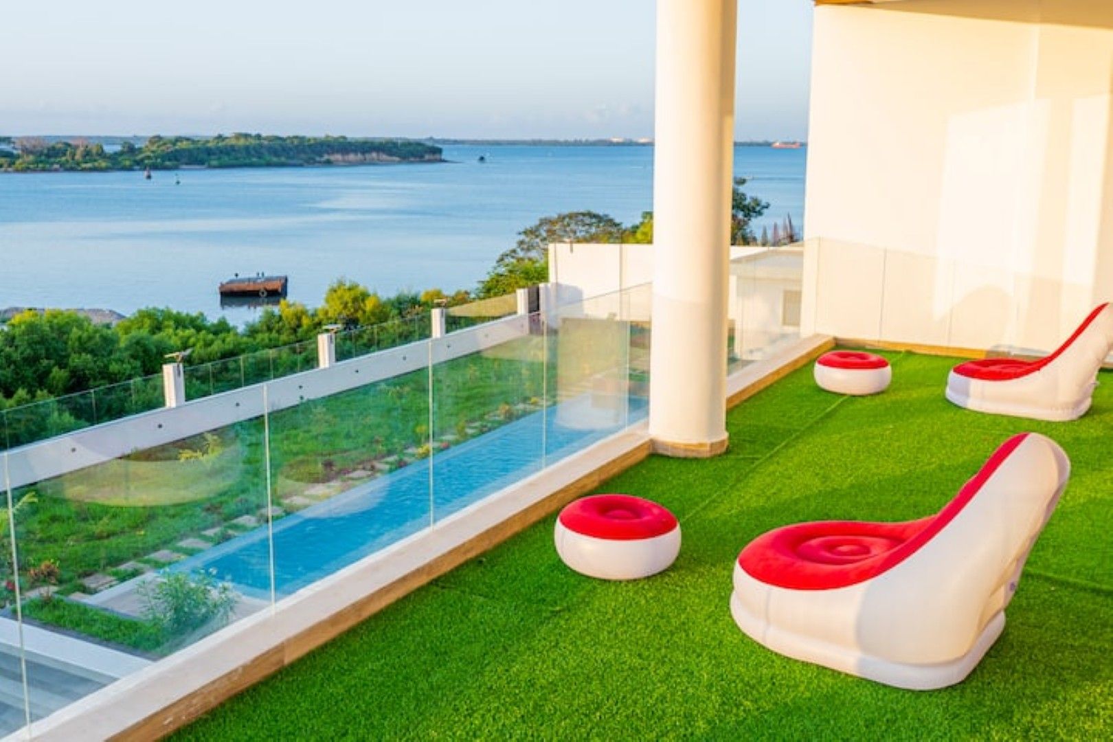 Ein Pool auf dem Dach mit Glasumrandung, grünem Kunstrasen und roten und weißen Liegestühlen.