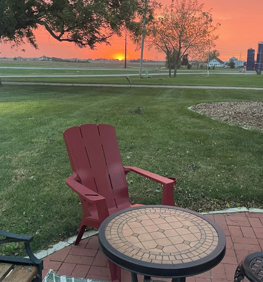Une chaise Adirondack rouge et une table ronde surmontée d’une mosaïque, installées sur une terrasse en briques, avec un champ verdoyant et un coucher de soleil en arrière-plan.