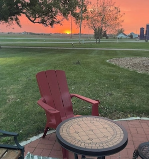 A red Adirondack chair and a round table with a mosaic top, set on a brick patio with a grassy field and a sunset in the background.