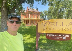 A man standing in front of a sign for Pella Mansion, which is a historic building with a large brick facade and arched windows.