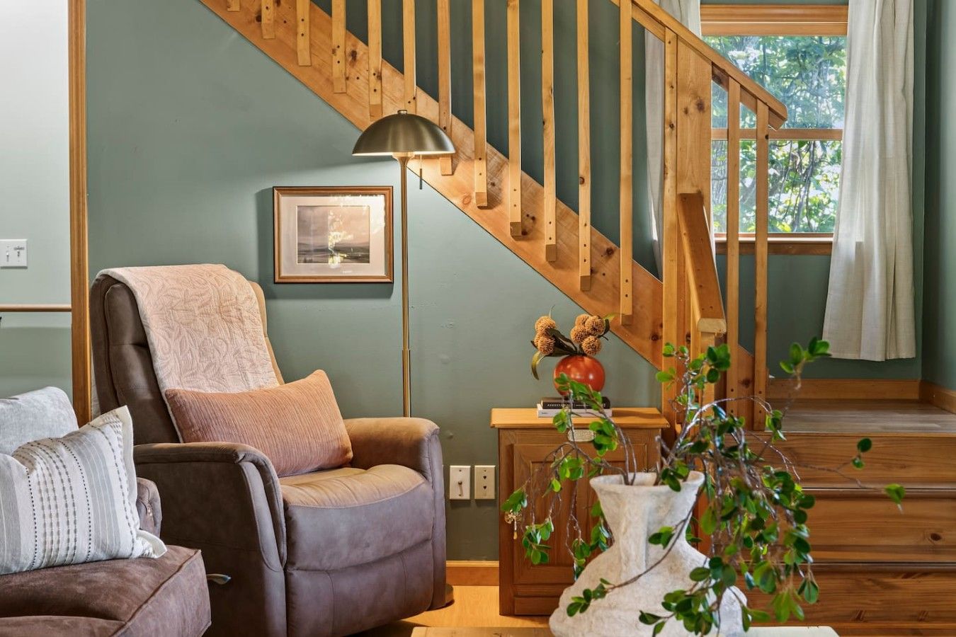 A living room with a wooden staircase, a leather armchair, a floor lamp, and a vase with dried flowers.