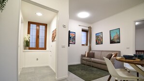 A living room with a sofa, a dining table, and a small side table. There are framed pictures on the wall and a window with curtains. - Fly&Rest MXP | Unit 1 | 24/7 Airport Transfer (Casorate Sempione)