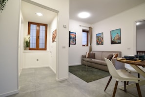 Een woonkamer met een bank, een eettafel en een klein bijzettafeltje. Er hangen foto's in lijsten aan de muur en een raam met gordijnen.