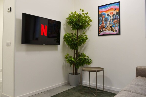 A living room with a flat-screen TV displaying the Netflix logo, a potted plant, a small round table, and a colorful poster on the wall.