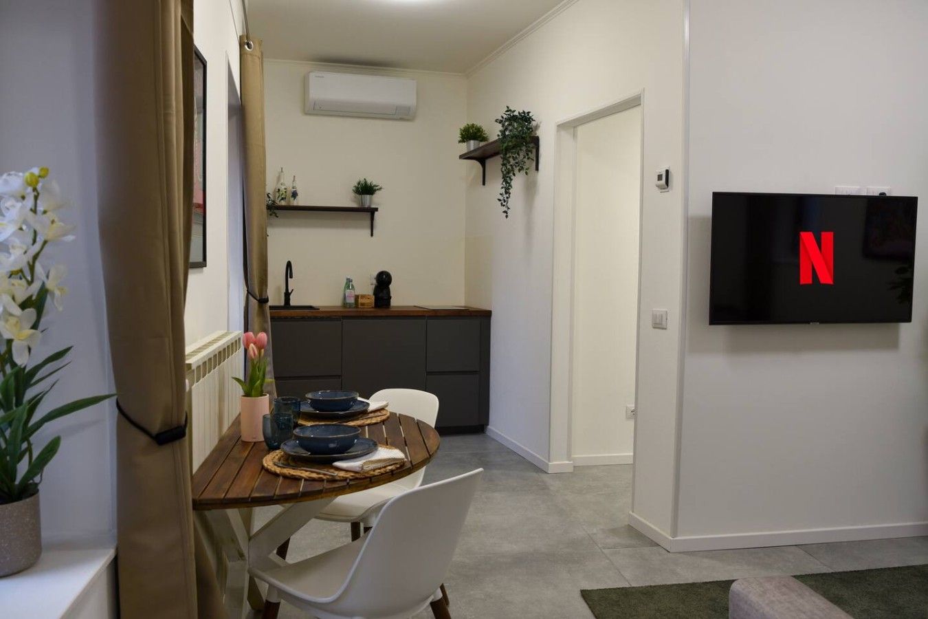 A modern kitchen with a dining area, a TV mounted on the wall, and a potted plant.