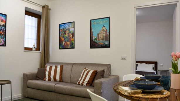 A living room with a sofa, a dining table with bowls, and two framed pictures on the wall. - Fly&Rest MXP | Unit 1 | 24/7 Airport Transfer (Casorate Sempione)
