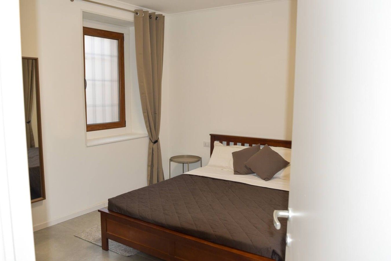 Une chambre à coucher avec un lit, une fenêtre avec des rideaux, une petite table ronde et une porte.