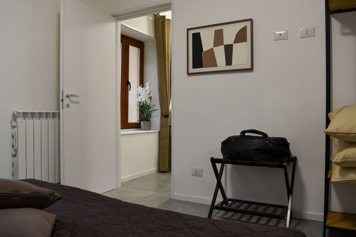Une chambre d’hôtel comprenant un lit, un radiateur, une fenêtre avec des rideaux, une œuvre d’art accrochée au mur, un porte-bagages et une étagère avec des oreillers.