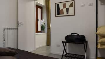 Une chambre d’hôtel comprenant un lit, un radiateur, une fenêtre avec des rideaux, une œuvre d’art accrochée au mur, un porte-bagages et une étagère avec des oreillers.